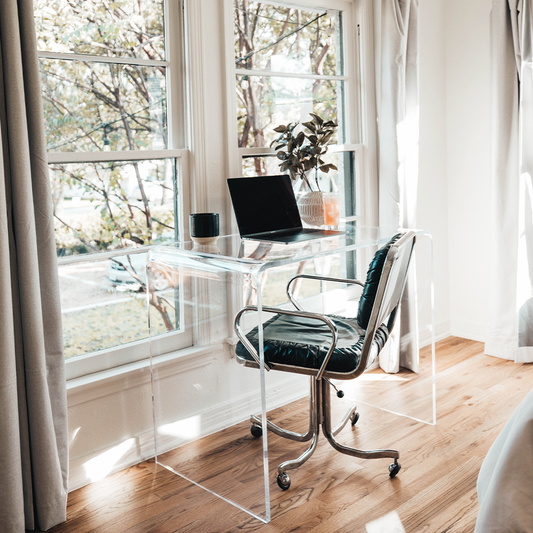 A clear acrylic waterfall edge vanity or desk displaying a laptop, a coffee cup, a plant, and a glass of juice placed in front of a large bedroom picture window with a chair.