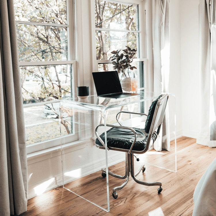 A clear acrylic waterfall desk in front of a window decorated with a candle, plant, glass of juice, and a laptop.