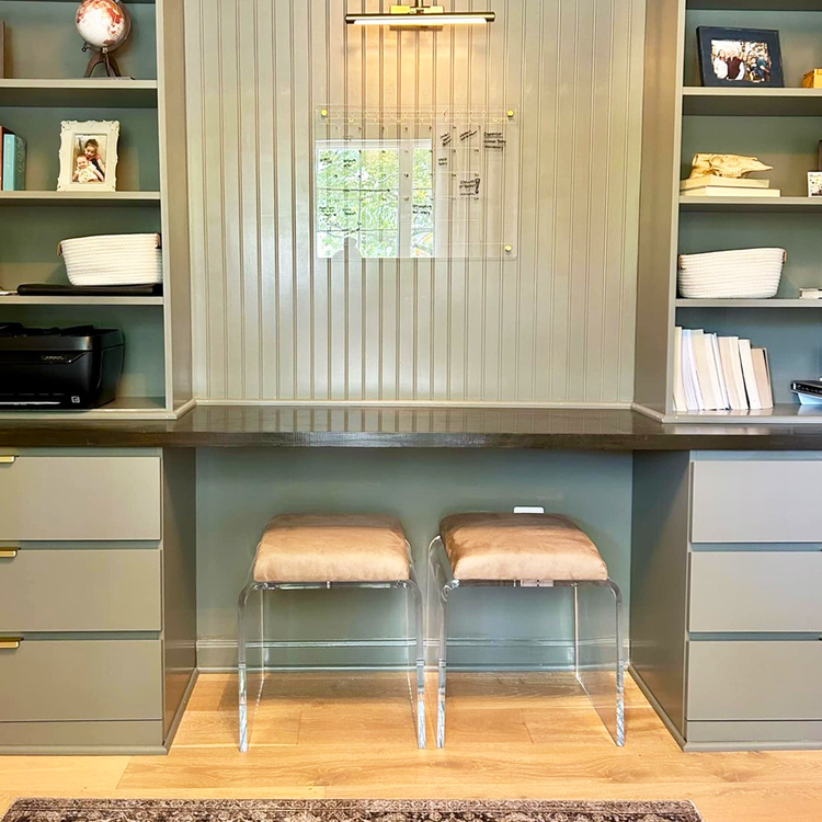 2 modern ottoman chairs with clear acrylic legs and light brown leather seat upholstery, placed underneath a counter/study area in a built-in wall bookshelf.
