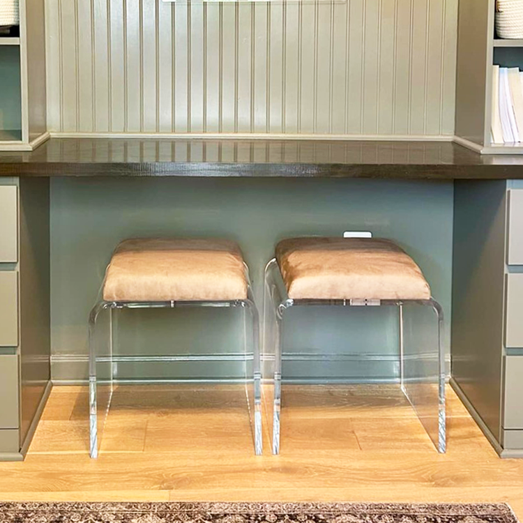 2 modern ottoman chairs with clear acrylic legs and light brown leather seat upholstery, placed underneath a counter/study area in a built-in wall bookshelf.