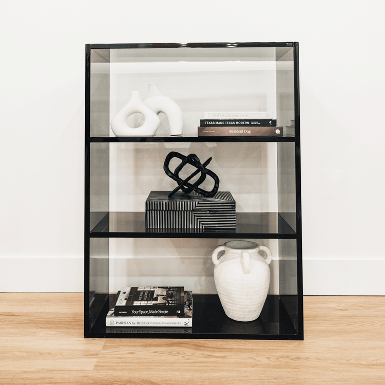 Clear gray tinted acrylic lucite bookshelf with 3 interior shelves displaying books and decor placed against a living room wall.