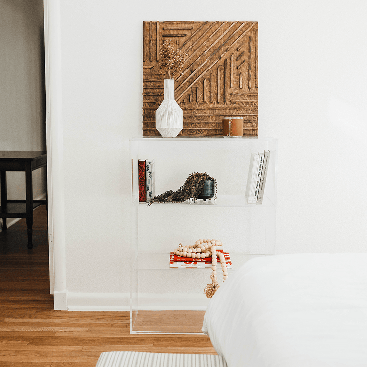 Clear acrylic lucite bookshelf with 3 interior shelves displaying books, plant and artwork in a bedroom.
