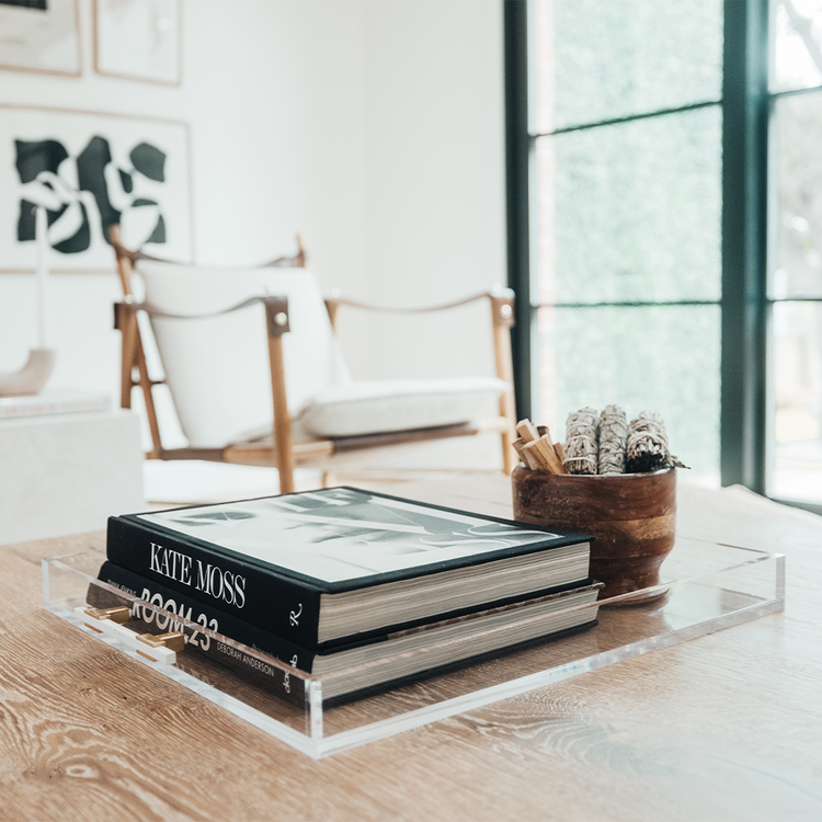 A clear acrylic lucite serving tray with gold and white handles holding decor and books placed on a wooden table.