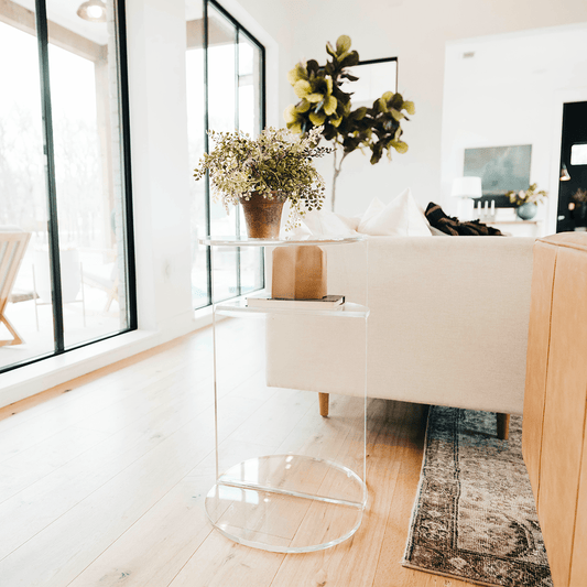 Clear acrylic side table featuring an extra shelf displaying a plant and decor beside a couch in a living room.