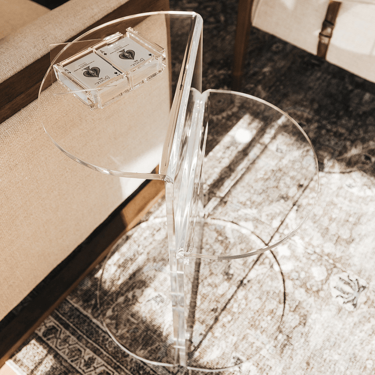 Close up of a clear acrylic side table featuring a top shelf extending 1 direction and the second shelf extending the opposite direction displaying a deck of cards in a clear acrylic card holder beside a chair.