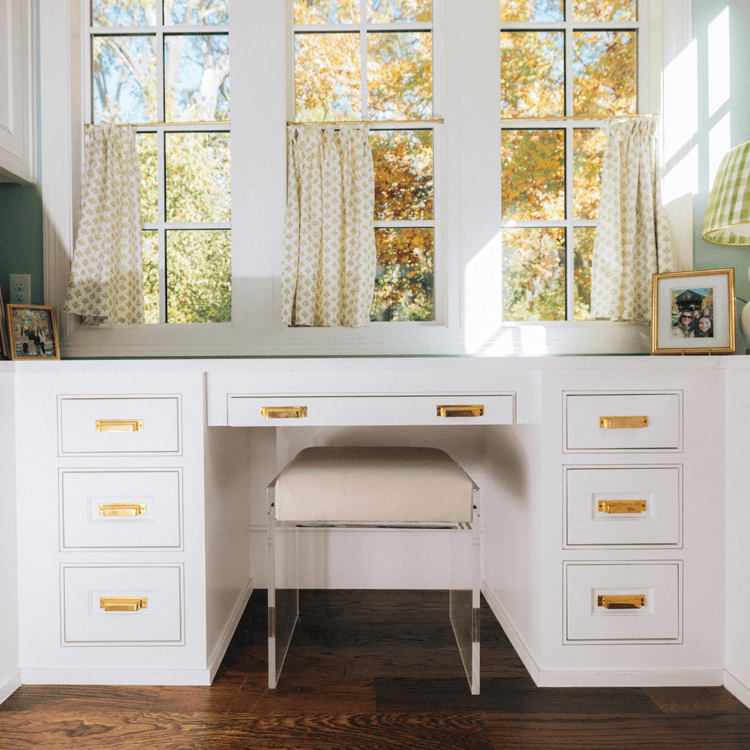 A clear acrylic accent chair with a beige upholstered seat slid underneath a white vanity with gold handles in a room with large windows and curtains.