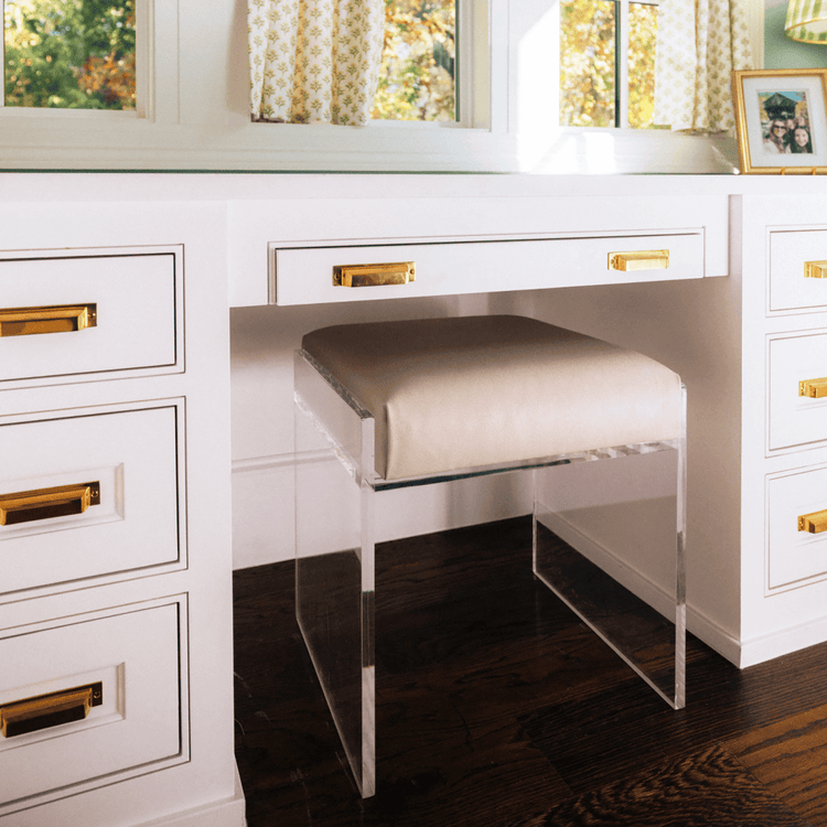 A clear acrylic accent chair with a beige upholstered seat slid underneath a white vanity with gold handles in a room with large windows and curtains.