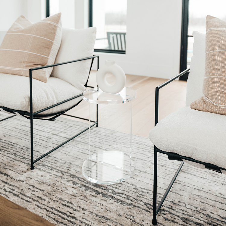 A clear acrylic lucite side table featuring a round top and round base displaying decor placed between 2 chairs in a living room.