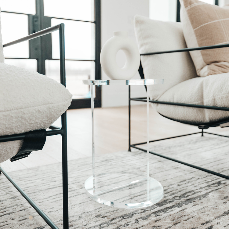 A clear acrylic lucite side table featuring a round top and round base displaying decor placed between 2 chairs in a living room.