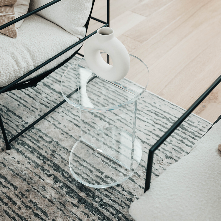 A clear acrylic lucite side table featuring a round top and round base displaying decor placed between 2 chairs in a living room.