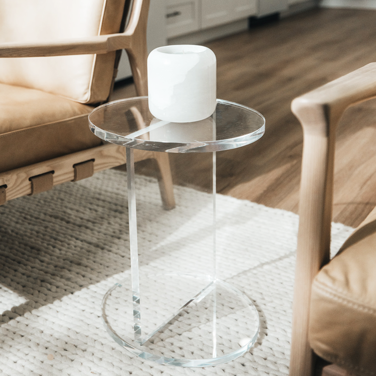 A clear acrylic lucite side table featuring a round top and round base displaying a candle placed between 2 chairs in a living room.