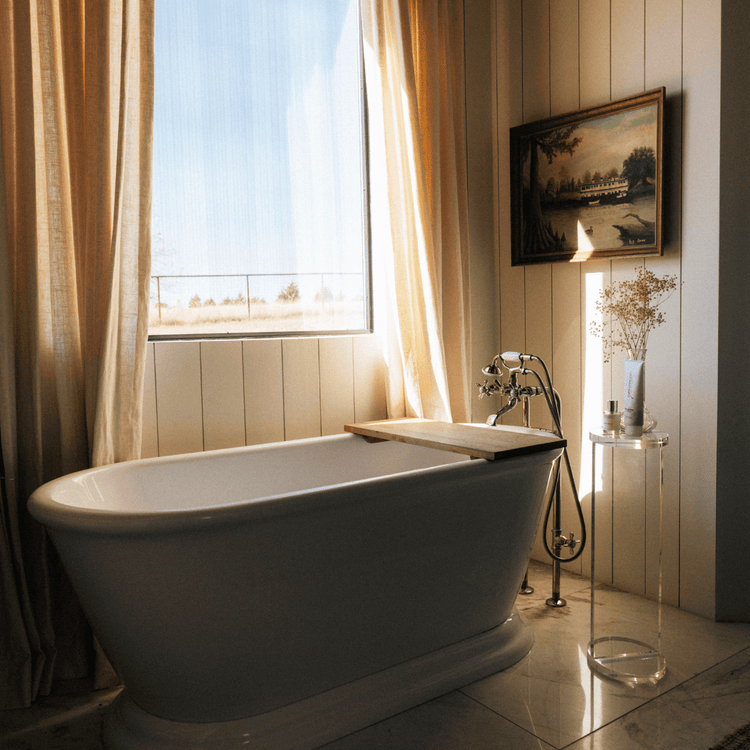 A clear acrylic lucite side table featuring a round top and round base displaying decor placed next to a soaking tub in a bathroom.