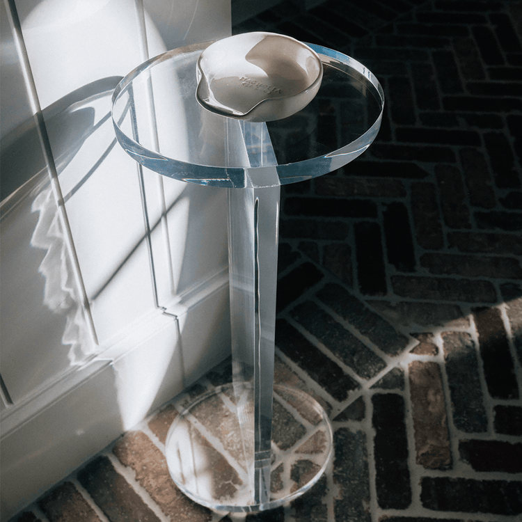 A clear acrylic lucite side table featuring a round top and round base displaying decor placed against a wall on a brick floor.