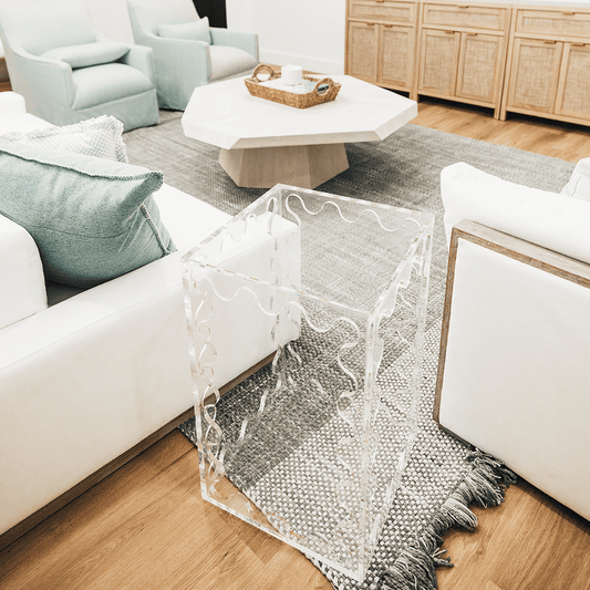 A clear acrylic lucite end table featuring wavy scalloped edges placed between a couch and a chair in a living room setting.