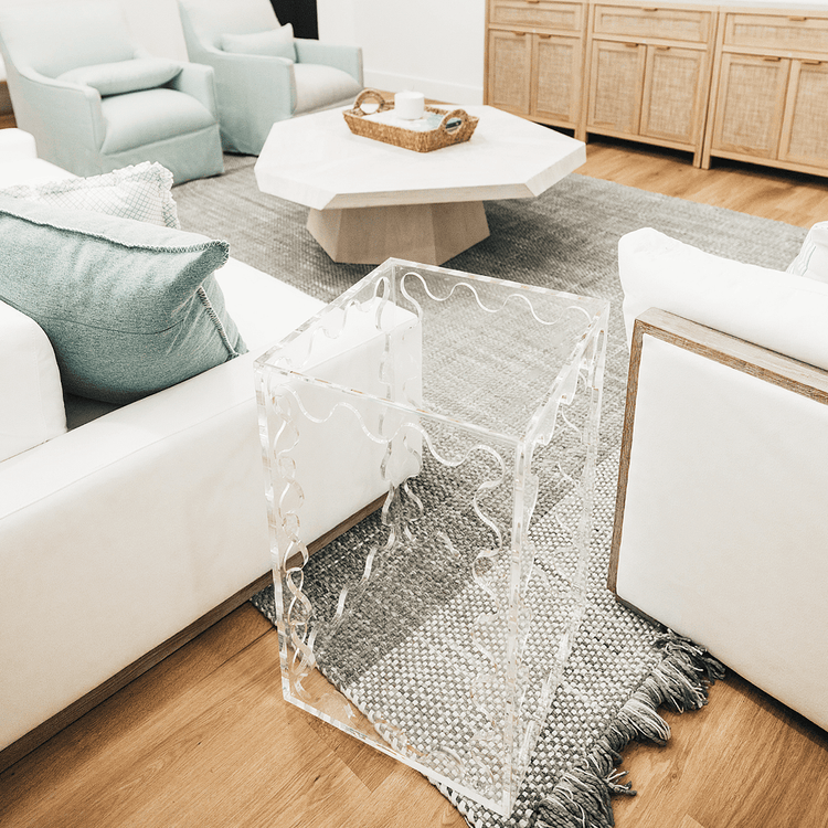 A clear acrylic lucite end table featuring wavy scalloped edges placed between a couch and a chair in a living room setting.