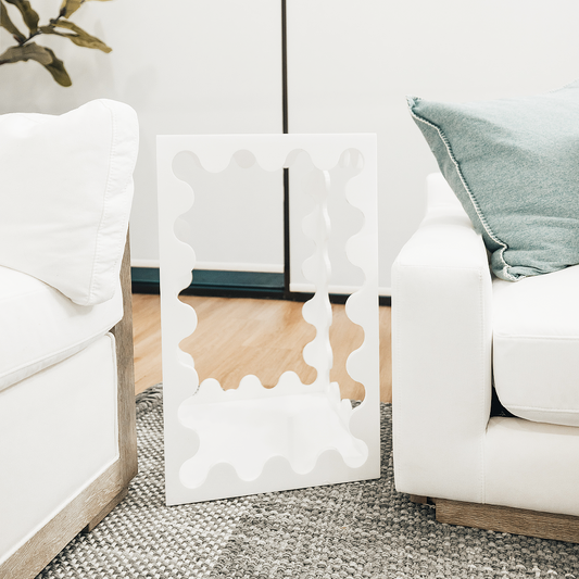 A white acrylic lucite end table featuring wavy scalloped edges placed between a couch and a chair in a living room setting.