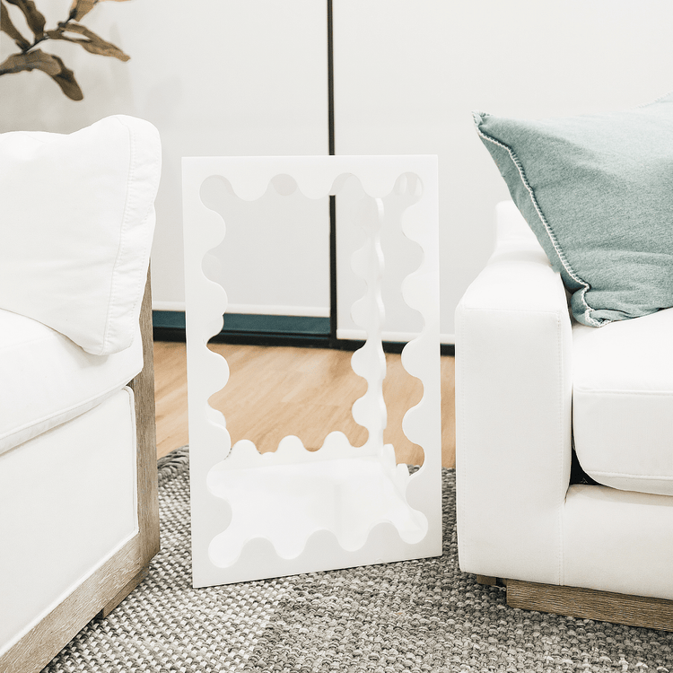 A white acrylic lucite end table featuring wavy scalloped edges placed between a couch and a chair in a living room setting.