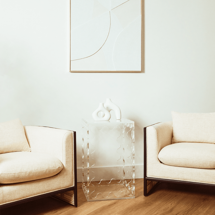 A clear acrylic lucite end table featuring wavy scalloped edges displaying ceramic decor placed between 2 easy chairs in a living room setting.