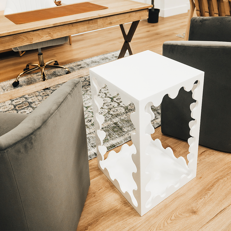 A white acrylic lucite end table featuring wavy scalloped edges placed between 2 easy chairs in an office setting.