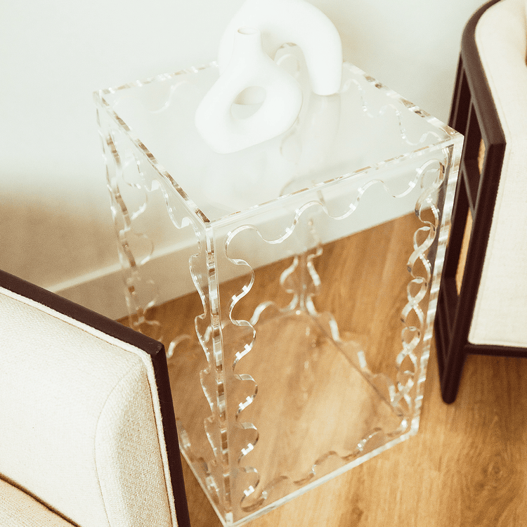 A clear acrylic lucite end table featuring wavy scalloped edges displaying ceramic decor placed between 2 easy chairs in a living room setting.