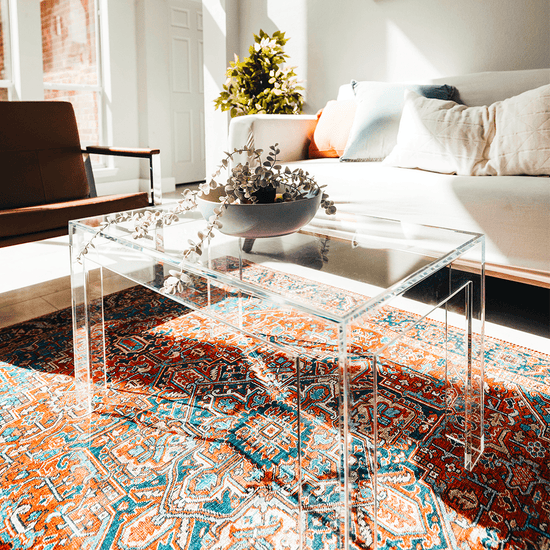 An acrylic lucite coffee table with square legs placed on a patterned rug, with a bowl of flowers on top.