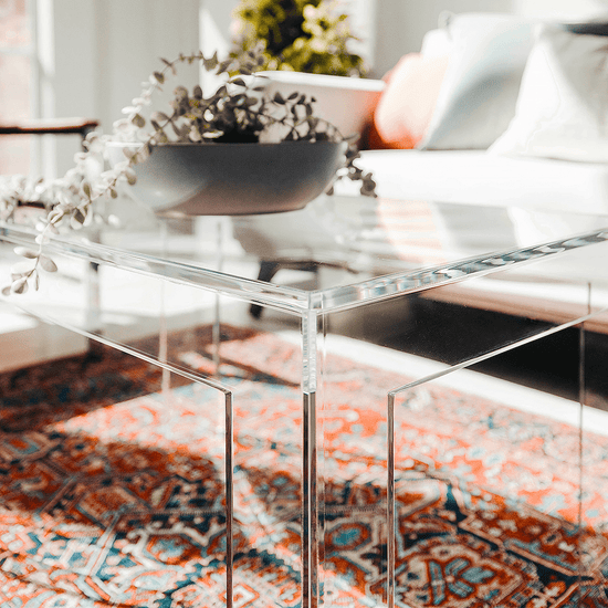 A closeup of an acrylic lucite coffee table with square legs placed on a patterned rug, with a bowl of flowers on top.