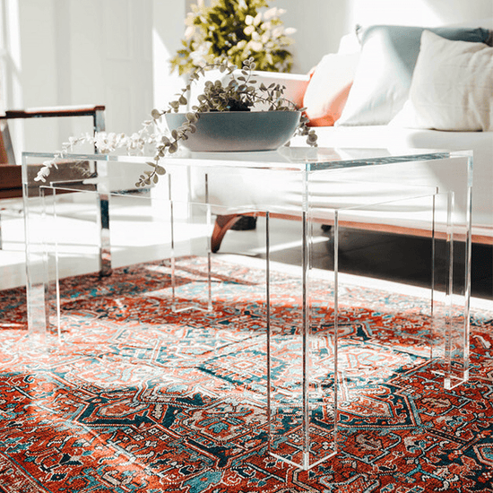 An acrylic lucite coffee table with square legs placed on a patterned rug, with a bowl of flowers on top.
