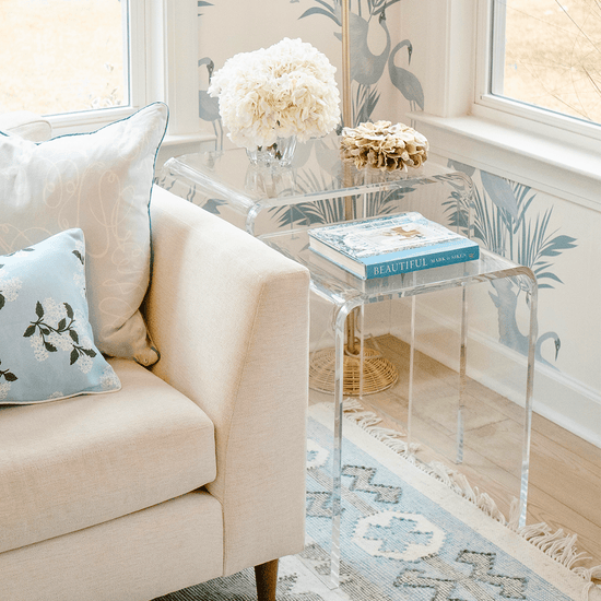 A clear acrylic nesting end table set placed next to a beige sofa with a decorative pillow on it, in a room with a floral wallpaper.