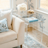 A clear acrylic nesting end table set placed next to a beige sofa with a decorative pillow on it, in a room with a floral wallpaper.
