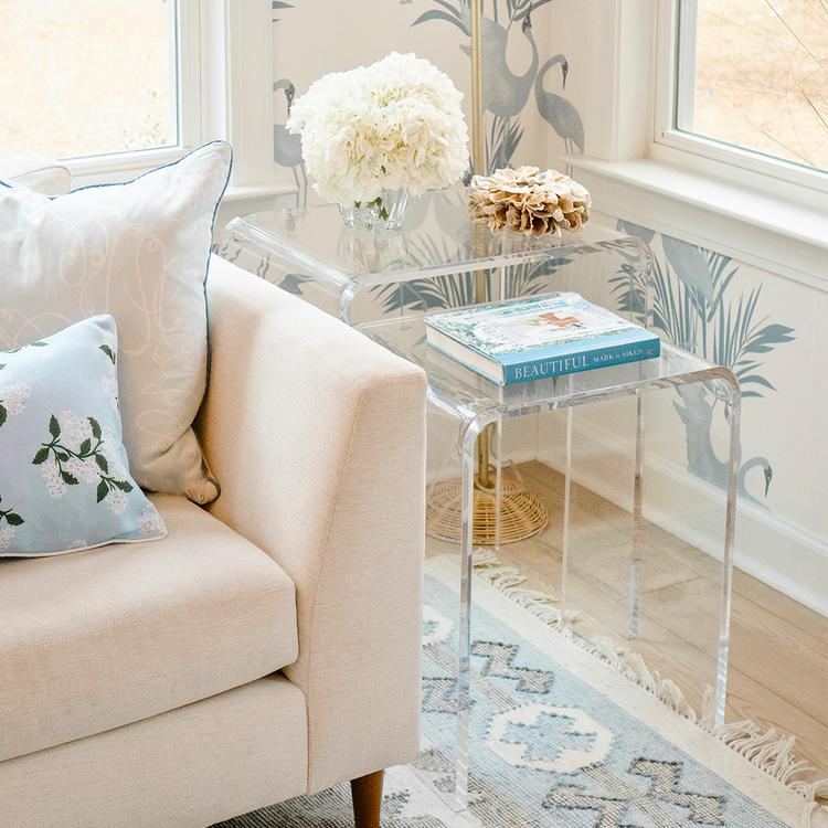 A clear acrylic nesting end table set placed next to a beige sofa with a decorative pillow on it, in a room with a floral wallpaper.