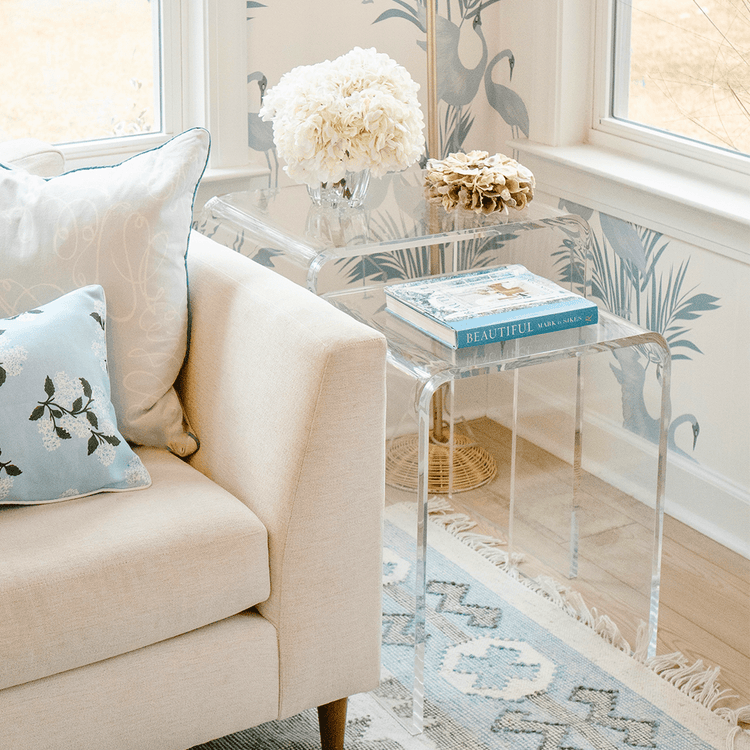 A clear acrylic nesting end table set placed next to a beige sofa with a decorative pillow on it, in a room with a floral wallpaper.
