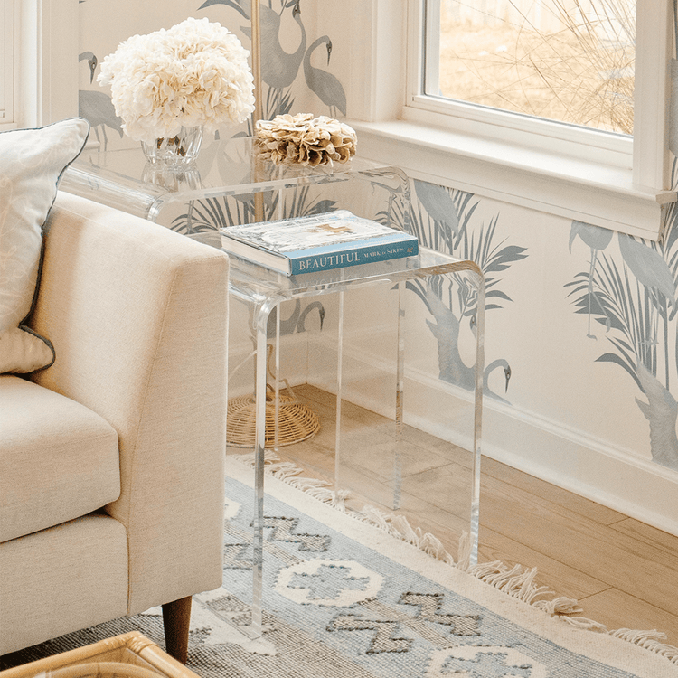 A clear acrylic nesting end table set placed in a living room setting beside a white sofa and windows.