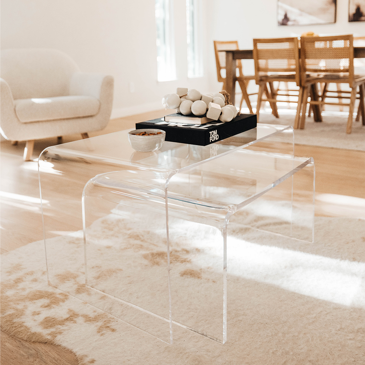A clear acrylic lucite nesting coffee table set with decor and books on top placed on a light white and brown cowhide rug in a living room setting.