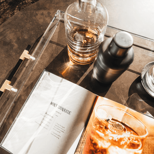 A clear acrylic lucite serving tray with gold and black handles holding a glass liquor carafe, glasses and drink receipe books placed on top of a coffee table.