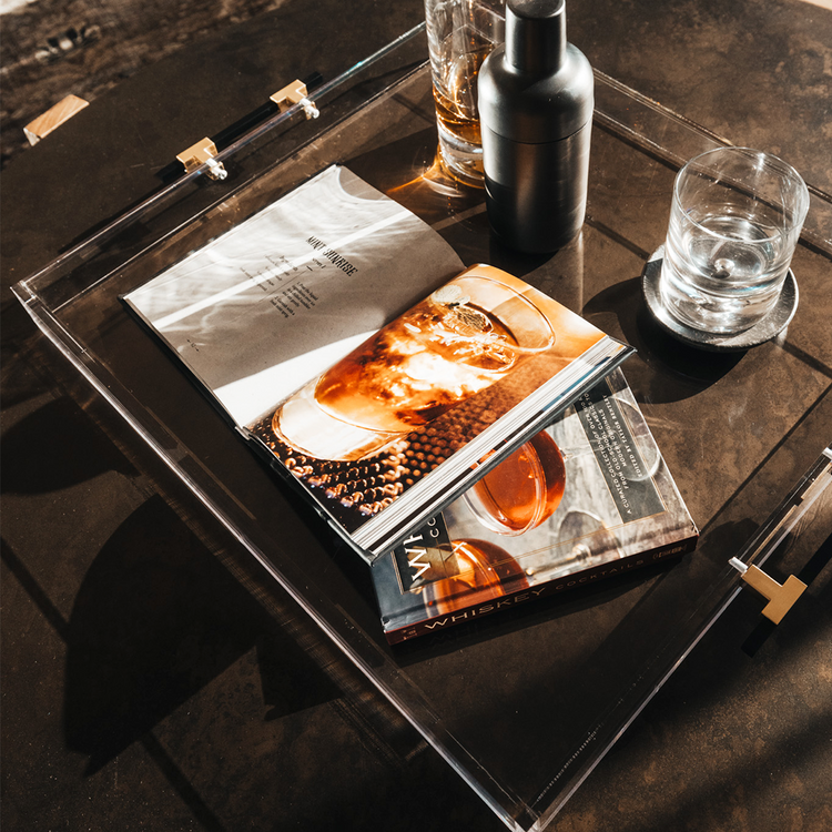 A clear acrylic lucite serving tray with gold and black handles holding a glass liquor carafe, glasses and drink receipe books placed on top of a coffee table.
