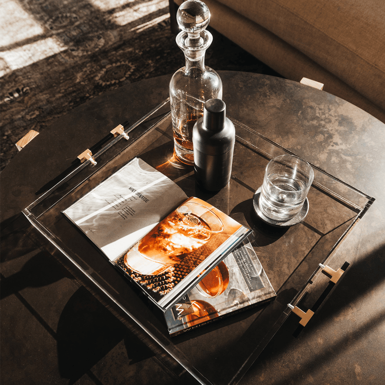 A clear acrylic lucite serving tray with gold and black handles holding a glass liquor carafe, glasses and drink receipe books placed on top of a coffee table.