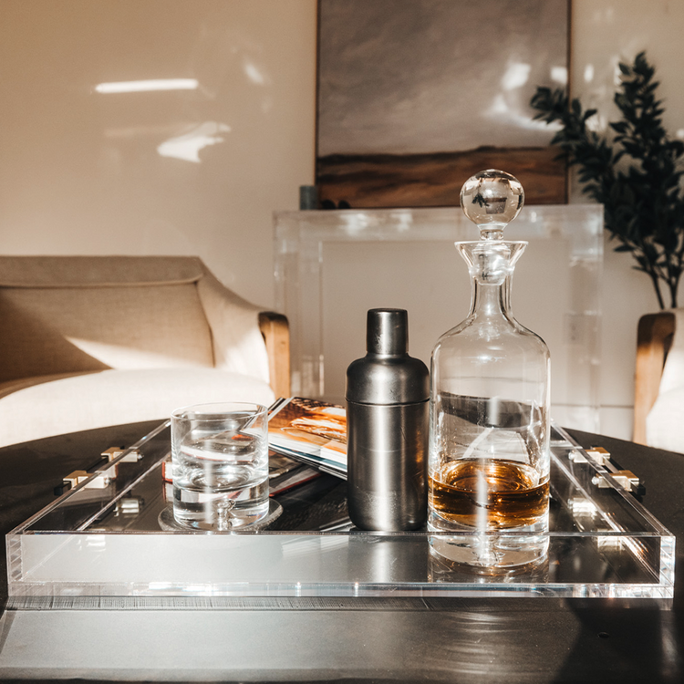 A clear acrylic lucite serving tray with gold and black handles holding a glass liquor carafe, glasses and drink receipe books placed on top of a coffee table.