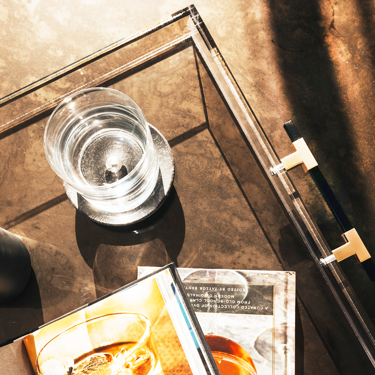 A close up of a clear acrylic lucite serving tray with gold and black handles holding a glass liquor carafe, glasses and drink receipe books placed on top of a coffee table.