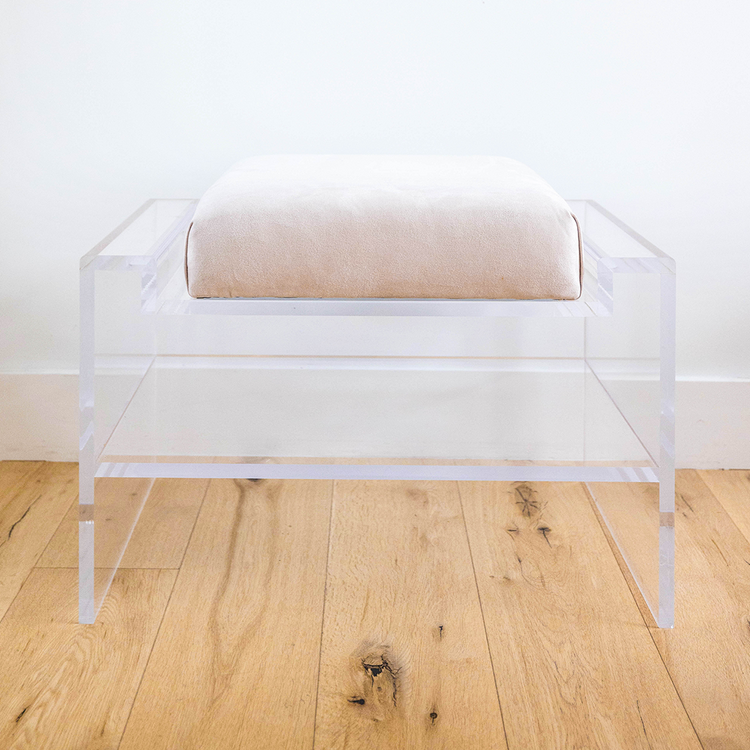 Catalog sample of a clear acrylic bench with an off-white cushion and storage shelf underneath on a wooden floor.