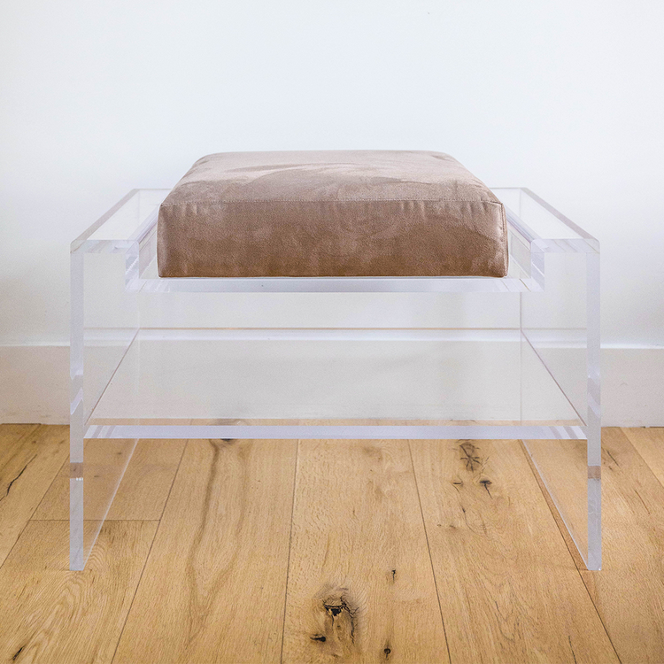 Catalog sample of a clear acrylic bench with a beige oat cushion and storage shelf underneath on a wooden floor.