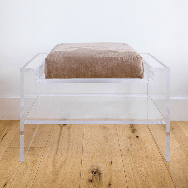 Catalog sample of a clear acrylic bench with a beige oat cushion and storage shelf underneath on a wooden floor.