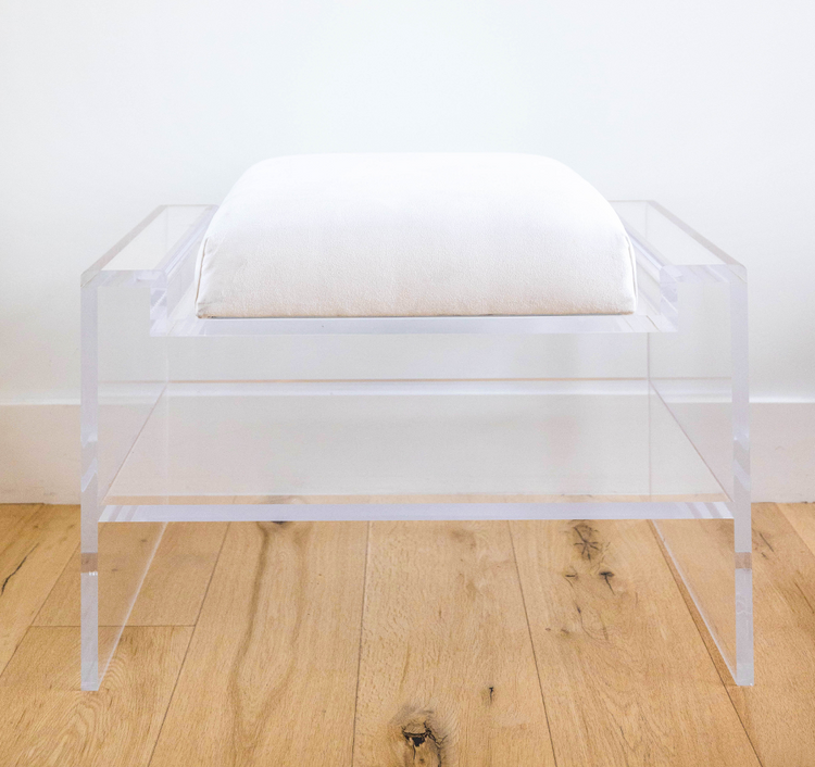 Catalog sample of a clear acrylic bench with a white cushion and storage shelf underneath on a wooden floor.