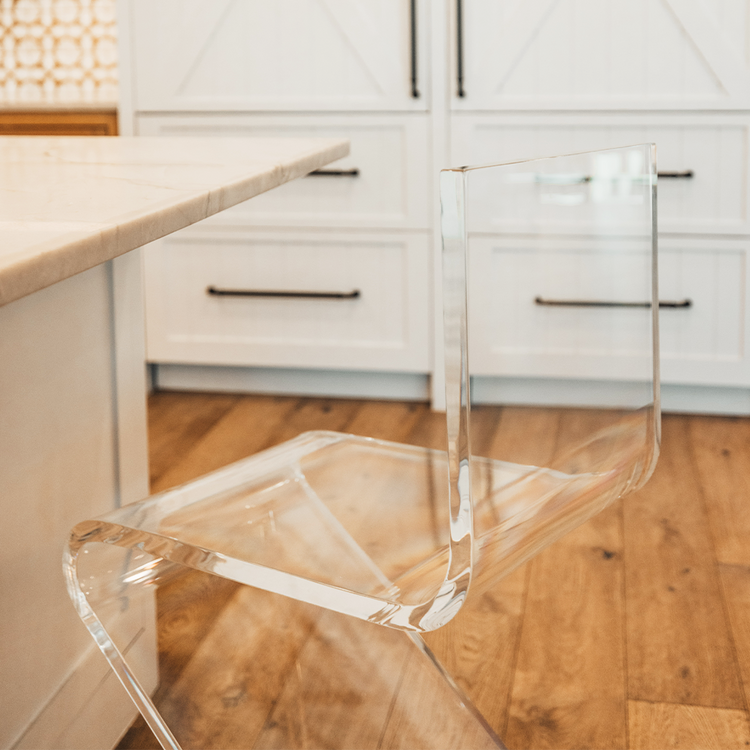 A close up of a clear acrylic bar stool with a modern design, placed in a kitchen setting next to a white countertop.