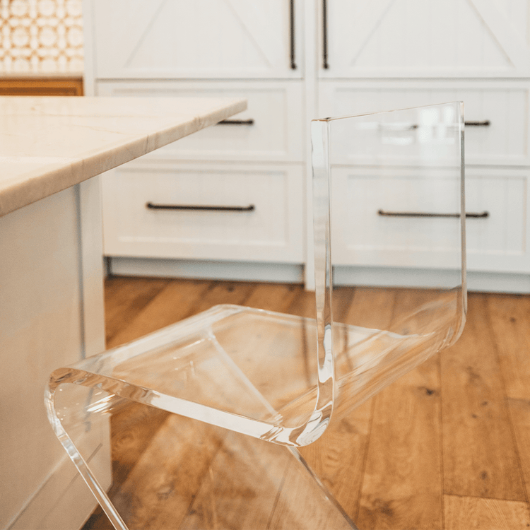 A close up of a clear acrylic bar stool with a modern design, placed in a kitchen setting next to a white countertop.