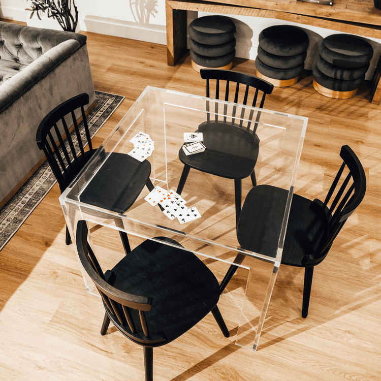 A top view of a square clear acrylic lucite game table featuring a deck of cards and a chair on each side of the table in a living room.