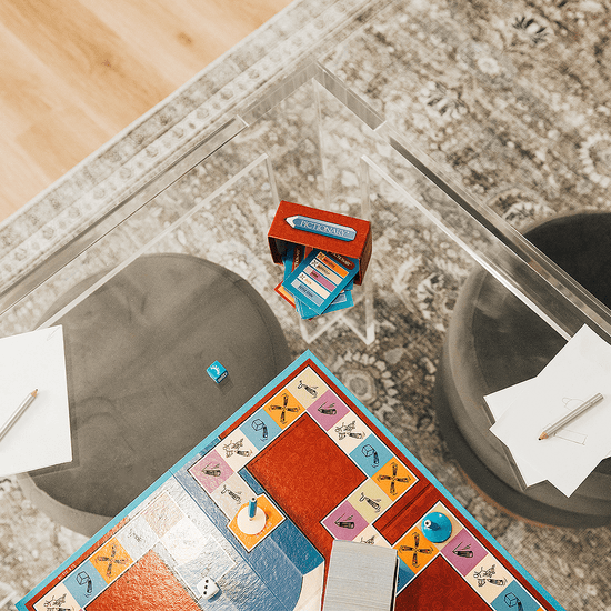 A closeup top view of a square clear acrylic lucite game table featuring a pictionary game and comfy padded stools on 2 sides of the table.