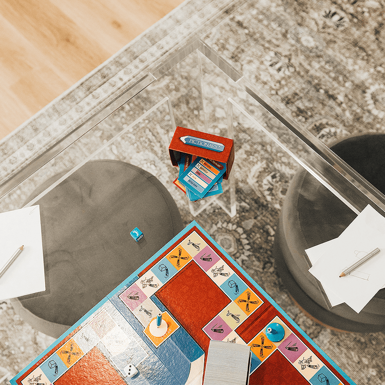 A closeup top view of a square clear acrylic lucite game table featuring a pictionary game and comfy padded stools on 2 sides of the table.