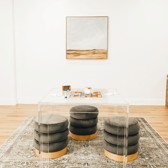 A closeup top view of a square clear acrylic lucite game table featuring a pictionary game and comfy padded stools on 3 sides of the table.
