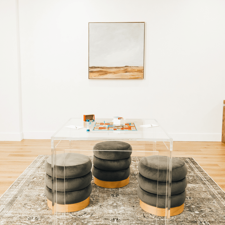 A closeup top view of a square clear acrylic lucite game table featuring a pictionary game and comfy padded stools on 3 sides of the table.