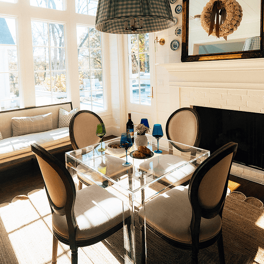 A square clear acrylic lucite game table displaying snacks and wine glasses with seating for 1 on each side in a brightly lit living room with lots of windows.